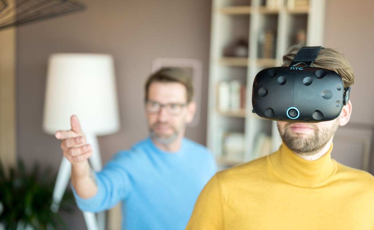 Ein Mann trägt ein VR-Headset, während ein anderer Mann im Hintergrund mit der Hand gestikuliert. Die Szene spielt sich in einem modernen Raum ab.