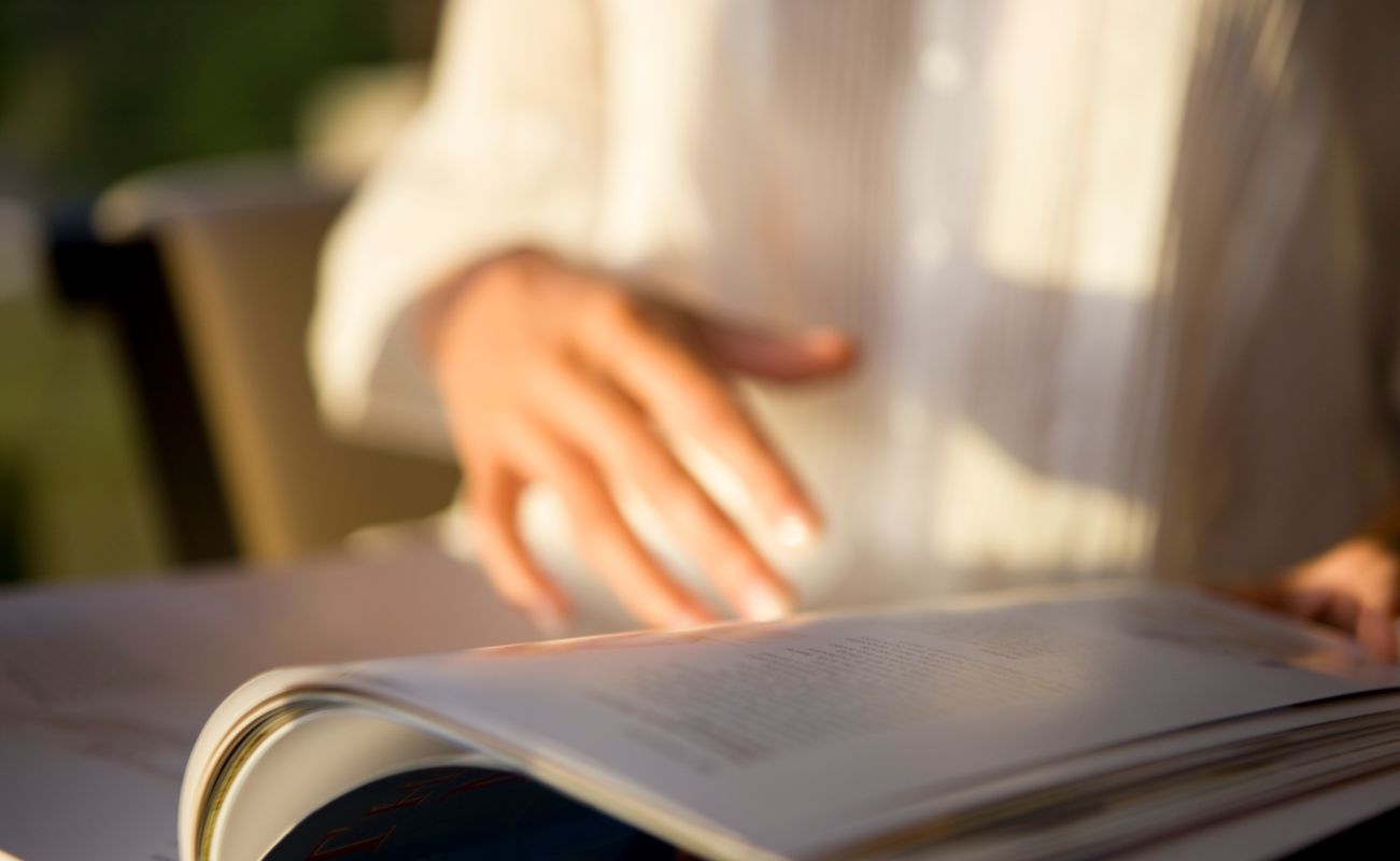 Eine Hand blättert durch eine Zeitschrift, die auf einem Tisch liegt. Weiches Licht fällt auf die Seite.